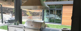 Modern outdoor kitchen with stainless steel grill and hood by ProlineRangeHoods.com, facing a guest house and lawn.