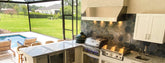 Outdoor kitchen by ProlineRangeHoods.com with grill, granite countertops, pool seating, and green backyard lawn.