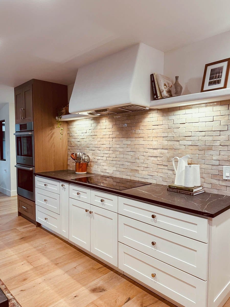 Modern kitchen with white cabinets, dark countertops, and brick backsplash; wooden floors and built-in oven. Stainless steel finish on appliances from ProlineRangeHoods.com adds a sleek touch.