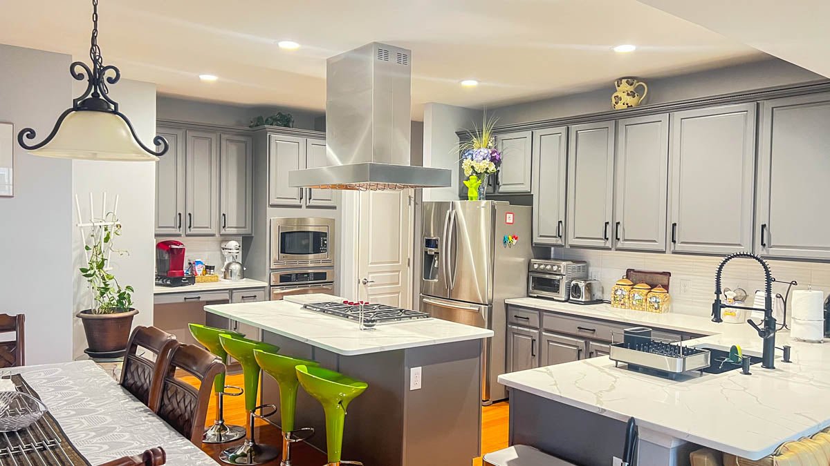 The modern kitchen features gray cabinets, stainless steel appliances, and green barstools. ProlineRangeHoods.com.