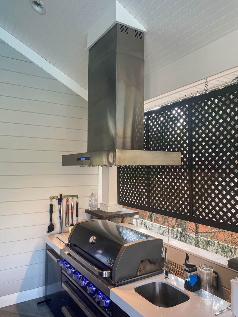 Outdoor kitchen with ProlineRangeHoods.com PLFI 755 island range hood, grill, & sink on white countertop under wood roof.