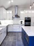 The modern kitchen features white cabinets, stainless appliances, gray tiles, and a blue island. ProlineRangeHoods.com ensures the efficiency.