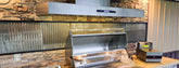 Stainless steel outdoor grill and vent hood by ProlineRangeHoods.com against a stone and corrugated metal wall.