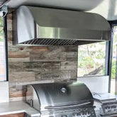 Stainless steel grill and vent hood by ProlineRangeHoods.com in an outdoor kitchen with wood-paneled walls and windows.