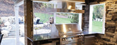 Modern outdoor kitchen with stainless steel grill, stone walls, large windows, and yard. ProlineRangeHoods.com.