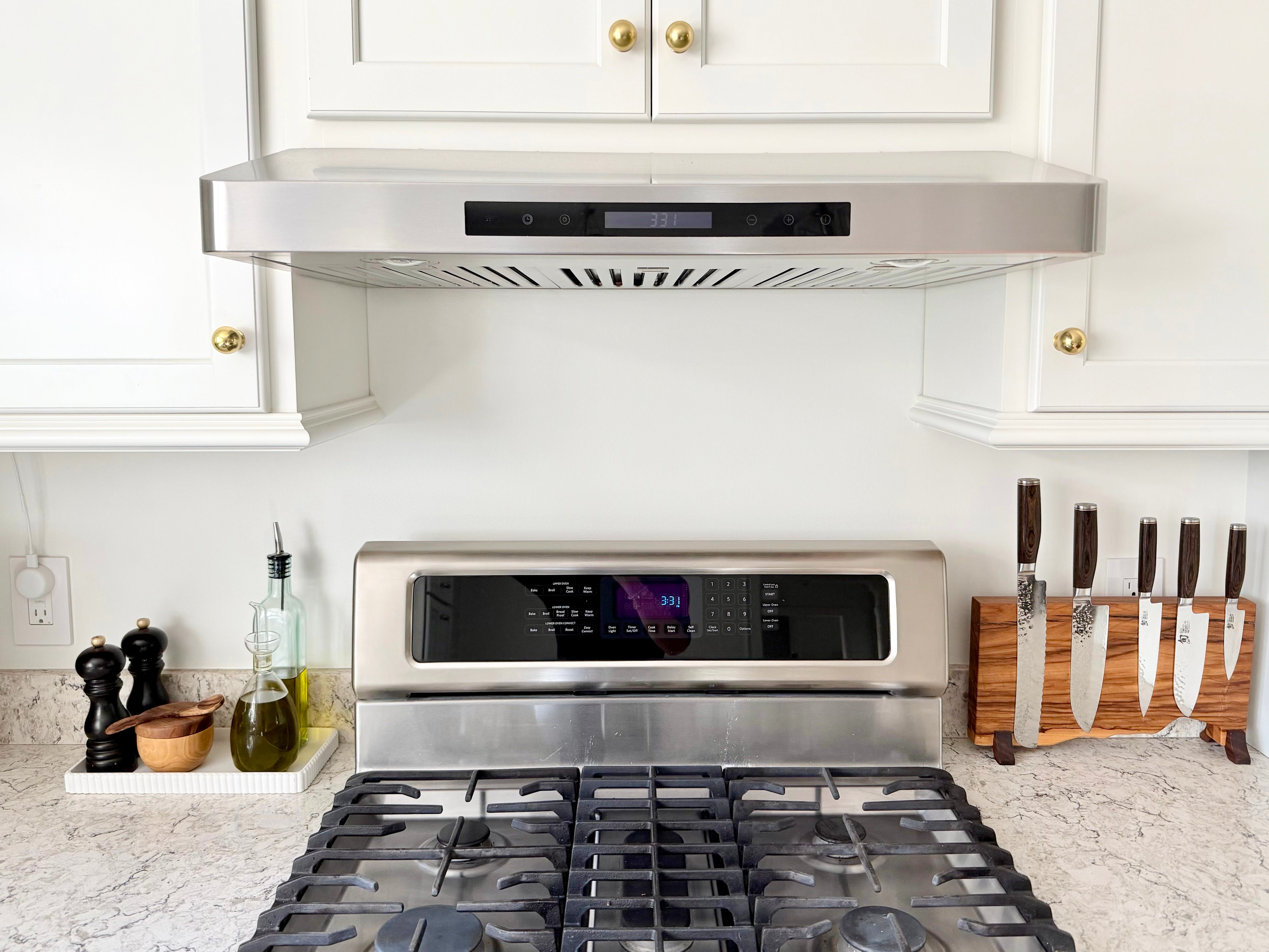 Modern kitchen stove with knives, spices, and olive oil on marble counter under white cabinets. ProlineRangeHoods.com.