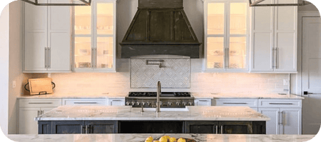 Modern kitchen with white cabinets, marble island, dark range hood by ProlineRangeHoods.com, and bowl of lemons.