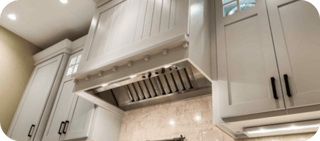 Modern white kitchen cabinets and a large stainless steel range hood above a tiled backsplash. ProlineRangeHoods.com.