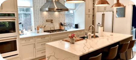 Modern kitchen with white cabinets, marble island, copper pendants, and stainless appliances. ProlineRangeHoods.com.