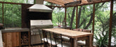 Outdoor kitchen with stainless steel grill, wooden dining table, chairs in a forest. ProlineRangeHoods.com.