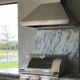 Stainless steel outdoor grill with vent hood on patio, grassy yard and pond view. ProlineRangeHoods.com.