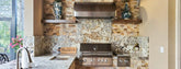 Outdoor kitchen with stainless grill, marble counters, and vases on shelves. ProlineRangeHoods.com.