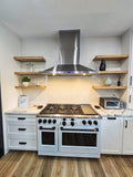 A contemporary kitchen with ProlineRangeHoods.com stainless steel stove, wooden shelves, and white cabinets on a wooden floor. PLFW 520.48