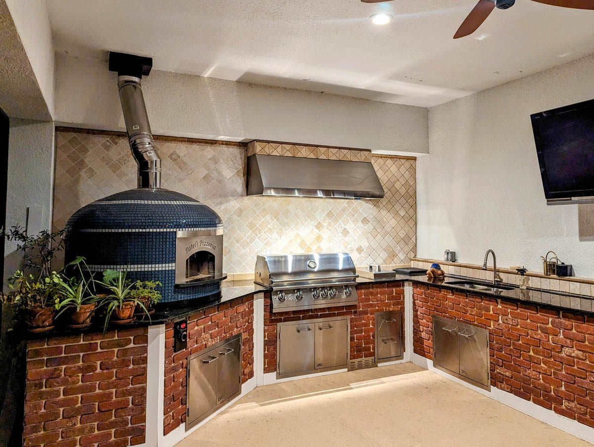 An outdoor kitchen with a ProlineRangeHoods.com PLJW 109 Hood features a pizza oven, grill, sink, and more for quiet performance.
