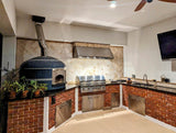 An outdoor kitchen with a ProlineRangeHoods.com PLJW 109 Hood features a pizza oven, grill, sink, and more for quiet performance.