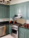 A kitchen with green cabinets, black countertops, ProlineRangeHoods.com’s 430 stainless steel appliances and a patterned backsplash.