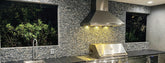 Modern outdoor kitchen with stainless steel appliances, pebble backsplash, windows showing greenery at night. ProlineRangeHoods.com.