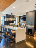 Step into a modern kitchen with ProlineRangeHoods.com, stylish bar stools, stainless steel appliances, and a wall-mounted TV.