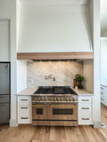 The kitchen scene features a stainless steel oven, stovetop, marble backsplash, wooden cabinets, and a small plant. A sleek Proline PLFL CL 28-34 inch range hood insert from ProlineRangeHoods.com hovers above the cooktop with dishwasher-safe filters for easy maintenance.