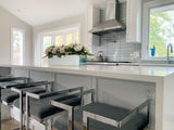 A modern kitchen with sleek bar stools and large windows features ProlineRangeHoods.com for an efficient ventilation system.