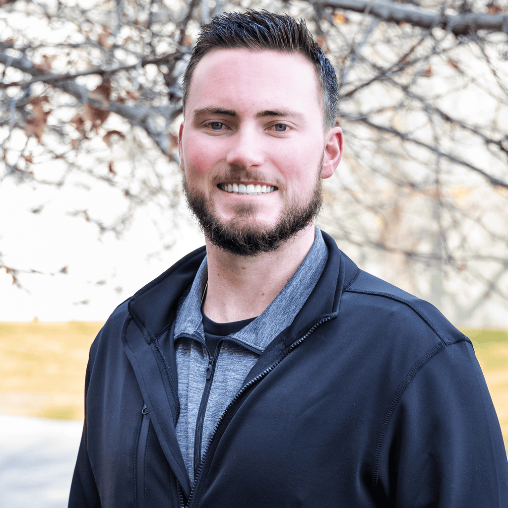 A man with short dark hair and a beard, wearing a black jacket and gray shirt, smiles outdoors. ProlineRangeHoods.com.