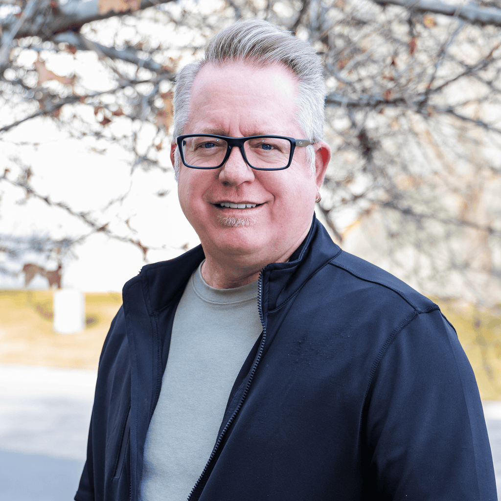Smiling man with glasses and gray hair, black jacket, outdoors by bare tree branches. ProlineRangeHoods.com.