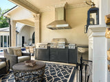 Outdoor kitchen with grill, black cabinets, patio set, rug, and pool, featuring a Proline PLGW 104 HE hood. ProlineRangeHoods.com.