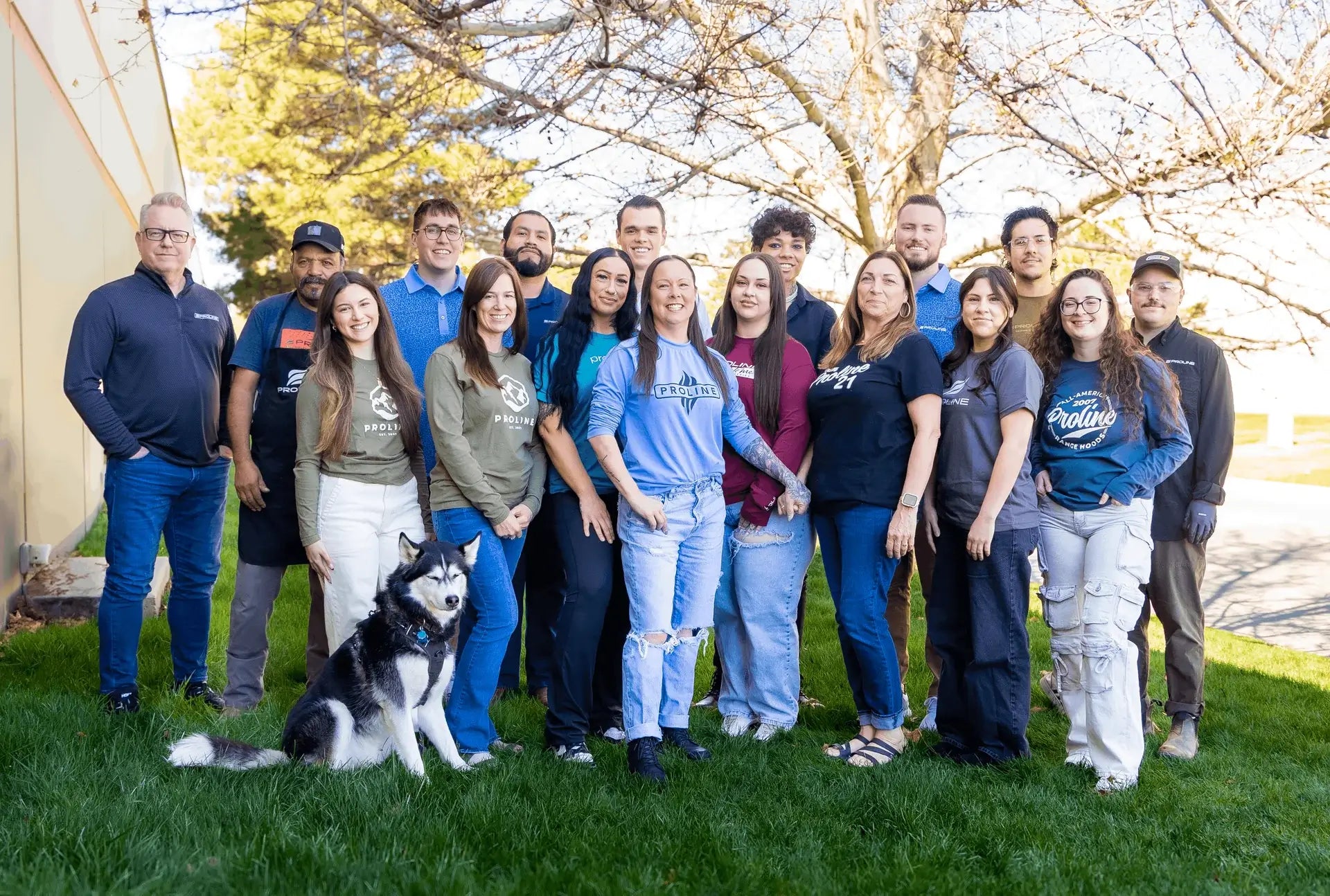 A group of people and a dog pose, smiling on grass with trees in the background. ProlineRangeHoods.com.