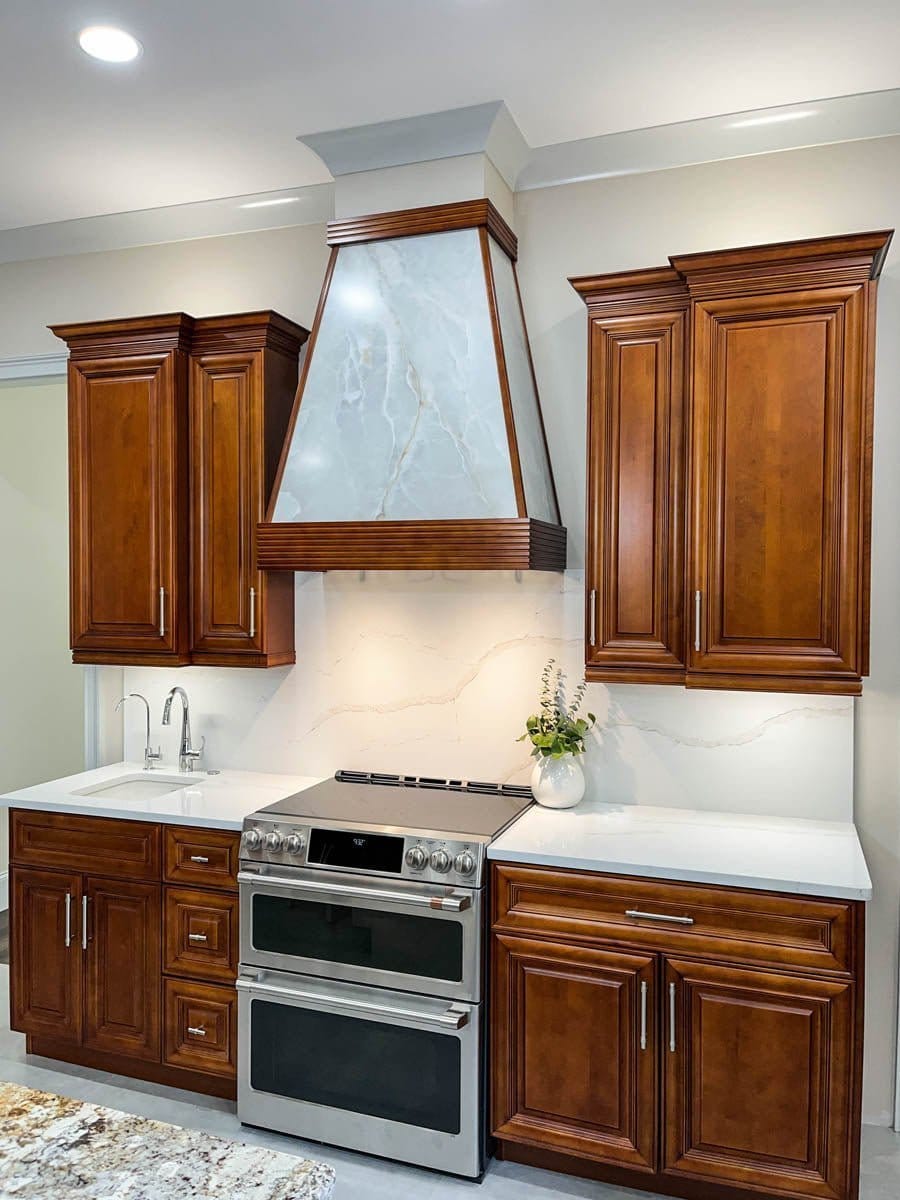 Modern kitchen with wooden cabinets, double oven, marble ProlineRangeHoods.com range hood (PLGL Classic, 1200 CFM), and vase.