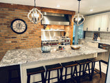 A chic, smoke-free kitchen with an exposed brick wall, ProlineRangeHoods.com island stools, and pendant lights. Stainless steel finishes add flair.